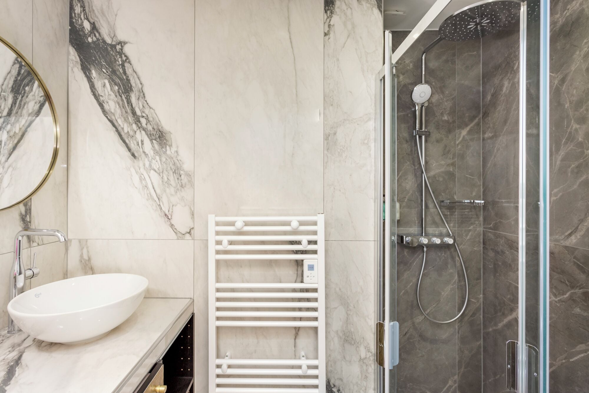 Un baño moderno con un lavamanos blanco, cabina de ducha con mampara de vidrio y pared con azulejos de mármol.