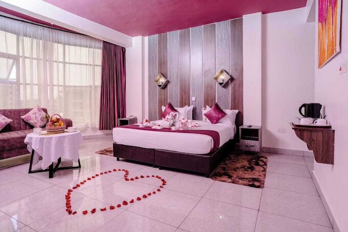 A modern bedroom with a large bed, a bedside table, a nightstand, a window with curtains, and a heart-shaped arrangement of rose petals on the floor.