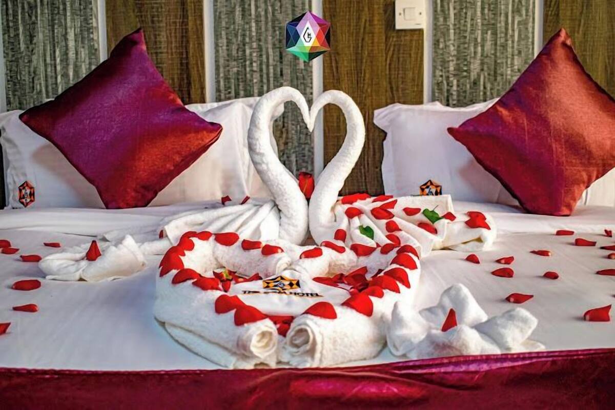 A bed with a heart-shaped towel arrangement and swans, red pillows, and a logo on the bedspread.