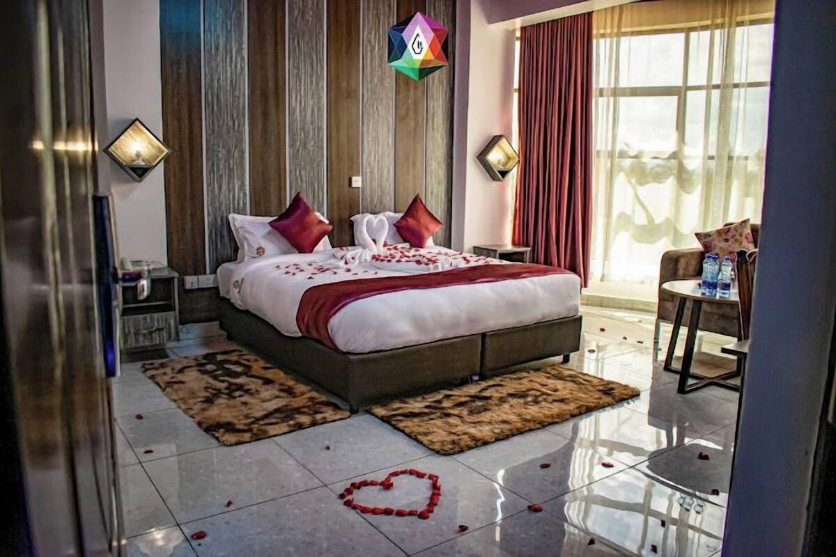 A modern hotel room with a large bed, wooden paneling, a view of greenery, and a small table with a chair.