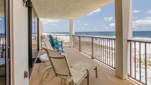 Un balcon avec deux chaises et une table, donnant sur une plage, avec une rambarde et une vue sur l’océan.