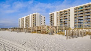 Un hôtel en bord de mer, avec plusieurs étages, une plage de sable en premier plan et une clôture en bois qui sépare la plage de l’hôtel.