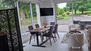 Un espace repas moderne avec une grande table et des chaises, un téléviseur fixé au mur et une vue sur l’extérieur grâce à de grandes fenêtres.