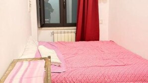 A small, single-bed room with a pink bedspread, a window with a red curtain, and a radiator below. - Max- Fortuna ground floor apartment (Ragusa)
