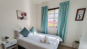 Una cama bien hecha con una colcha blanca y dos almohadas, una mesita de noche con una planta, una ventana con cortinas color turquesa y un cuadro en la pared.