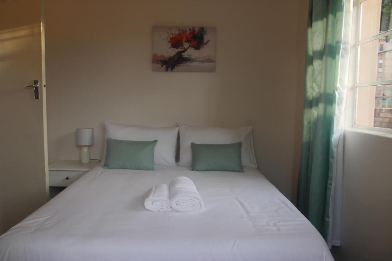 Een hotelkamer met een bed, twee kussens, een aan de muur hangend schilderij, een nachtkastje met een lamp en een raam met gordijnen.