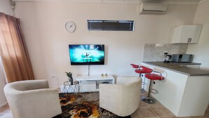 Un espacio habitable compacto con sofá, una pequeña mesa de comedor, un televisor y una zona de cocina con microondas, además de un taburete rojo.