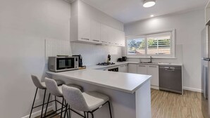A modern kitchen with white cabinetry, a built-in microwave, a gas stove, and a dishwasher. There are two bar stools and a window providing natural light. - Kennington House, 3BR Home in Bendigo City (Kennington)