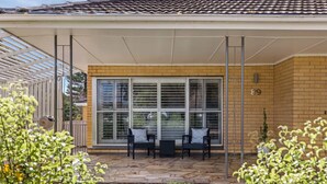 A covered patio with two black wicker chairs and a table, a large sliding glass door, and a brick wall. - Kennington House, 3BR Home in Bendigo City (Kennington)