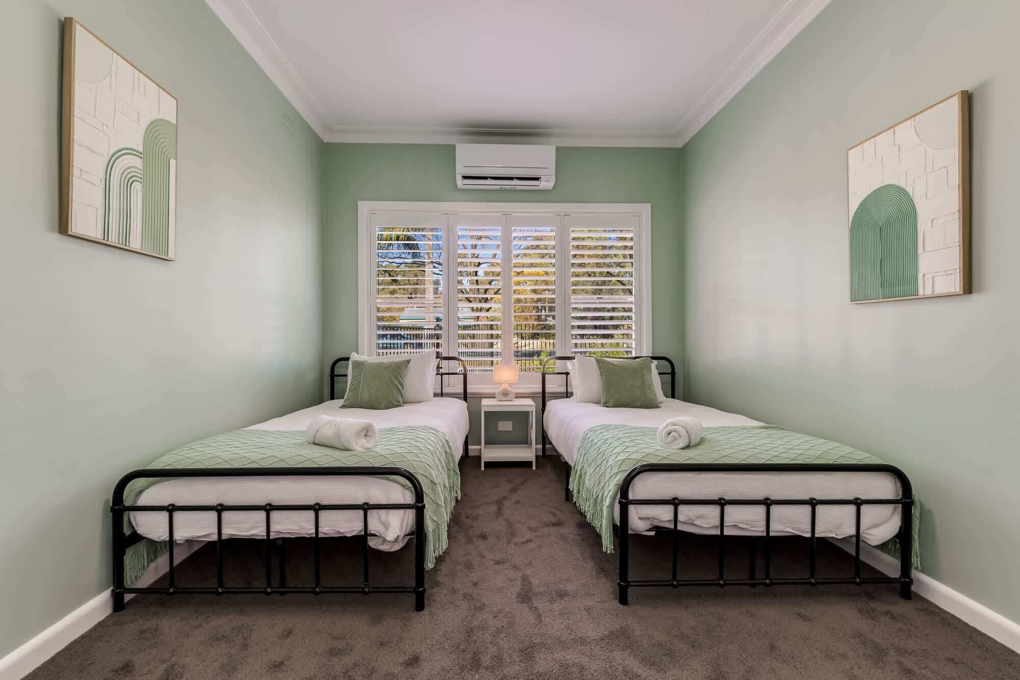 A room with two beds, a window with blinds, an air conditioning unit, and artwork on the walls.