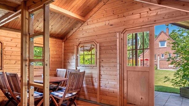 Een houten hut met een eethoek, grote ramen en uitzicht op een stenen huis.