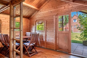 A wooden cabin with a dining area, large windows, and a view of a brick house. - Holiday Home 'Svea' with Private Terrace, Private Garden and Wi-Fi (Cuxhaven)