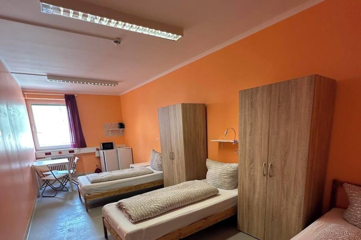 A dormitory room with three single beds, a wooden wardrobe, a desk with a chair, and a radiator.