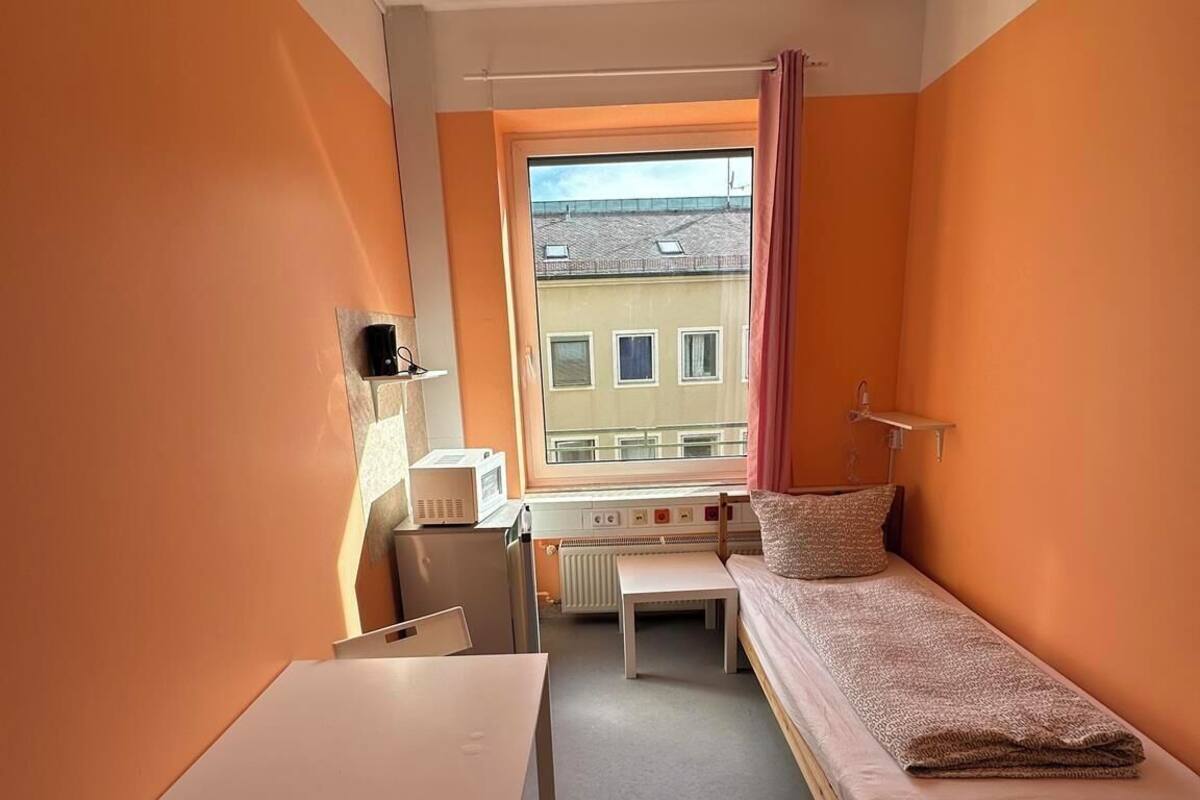 A compact room with a single bed, a small table, and a chair. A window provides a view of buildings outside.