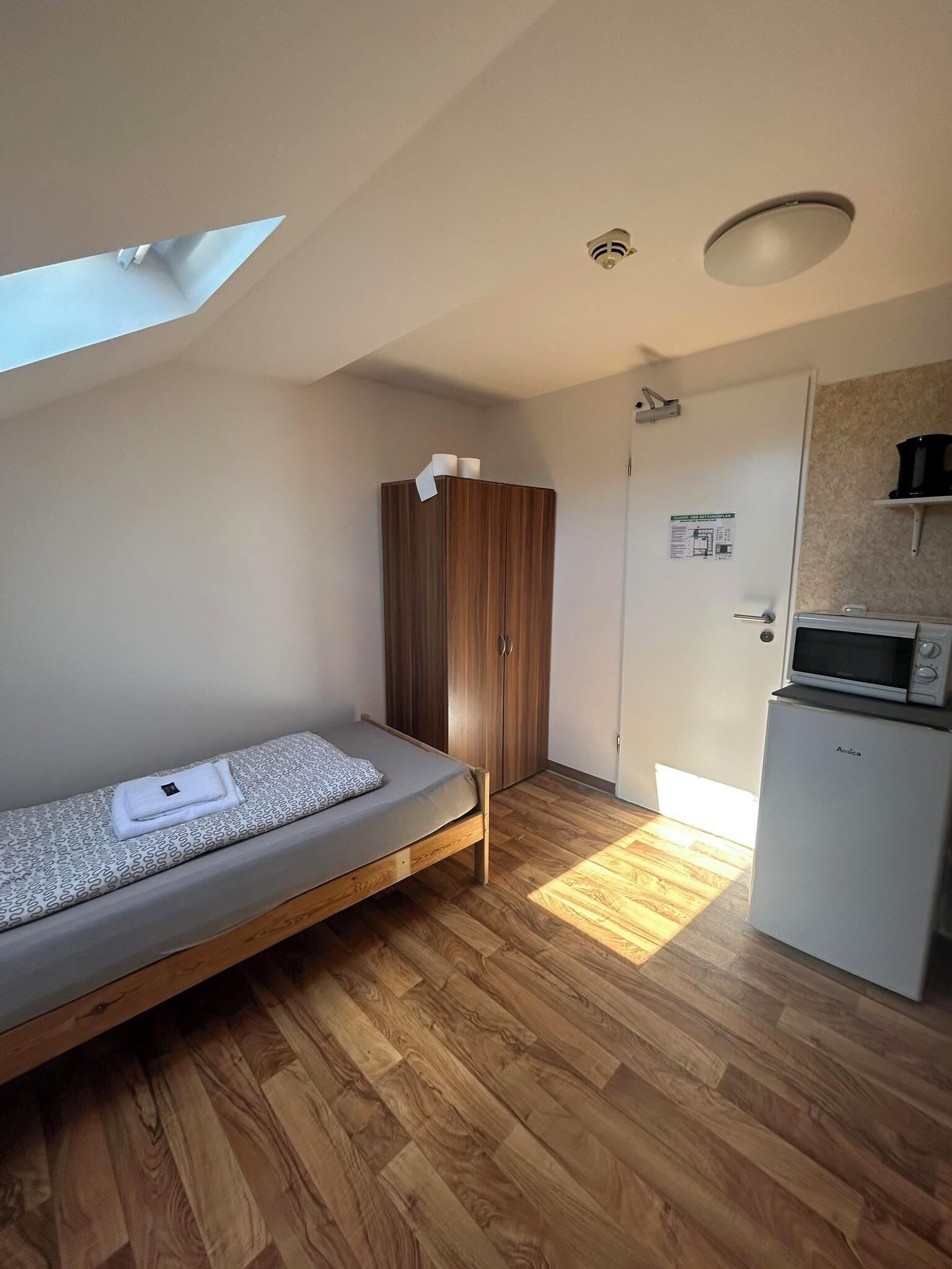 A compact room with a single bed, a wardrobe, a kitchenette, and a door with a notice.