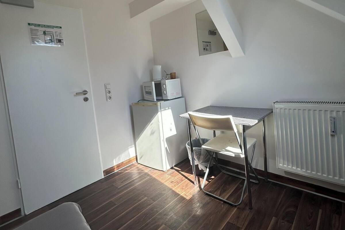 A room with a sloped ceiling, a skylight, a white radiator, a small white fridge, a white table with a chair, and a wooden floor.