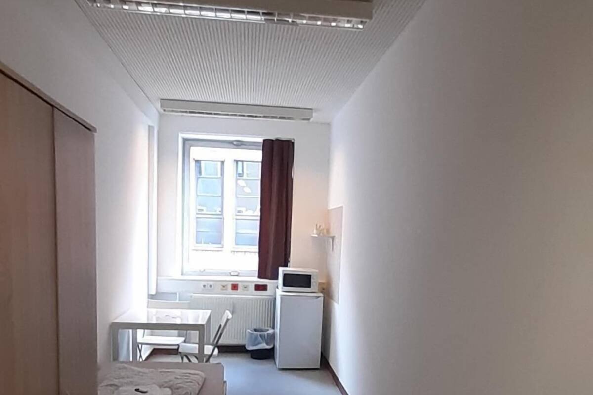 A narrow hallway with a bed, a desk, and a window with curtains.