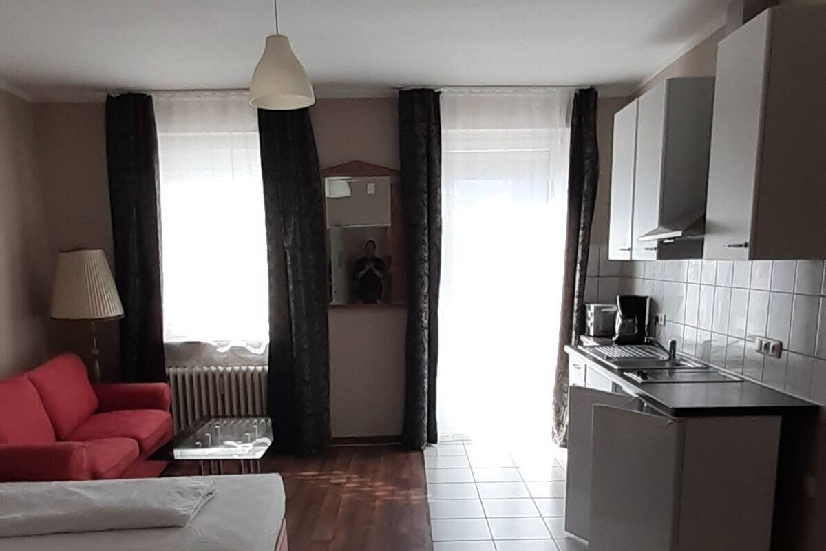 A compact hotel room with a bed, a red sofa, a kitchenette, and a view of the street.