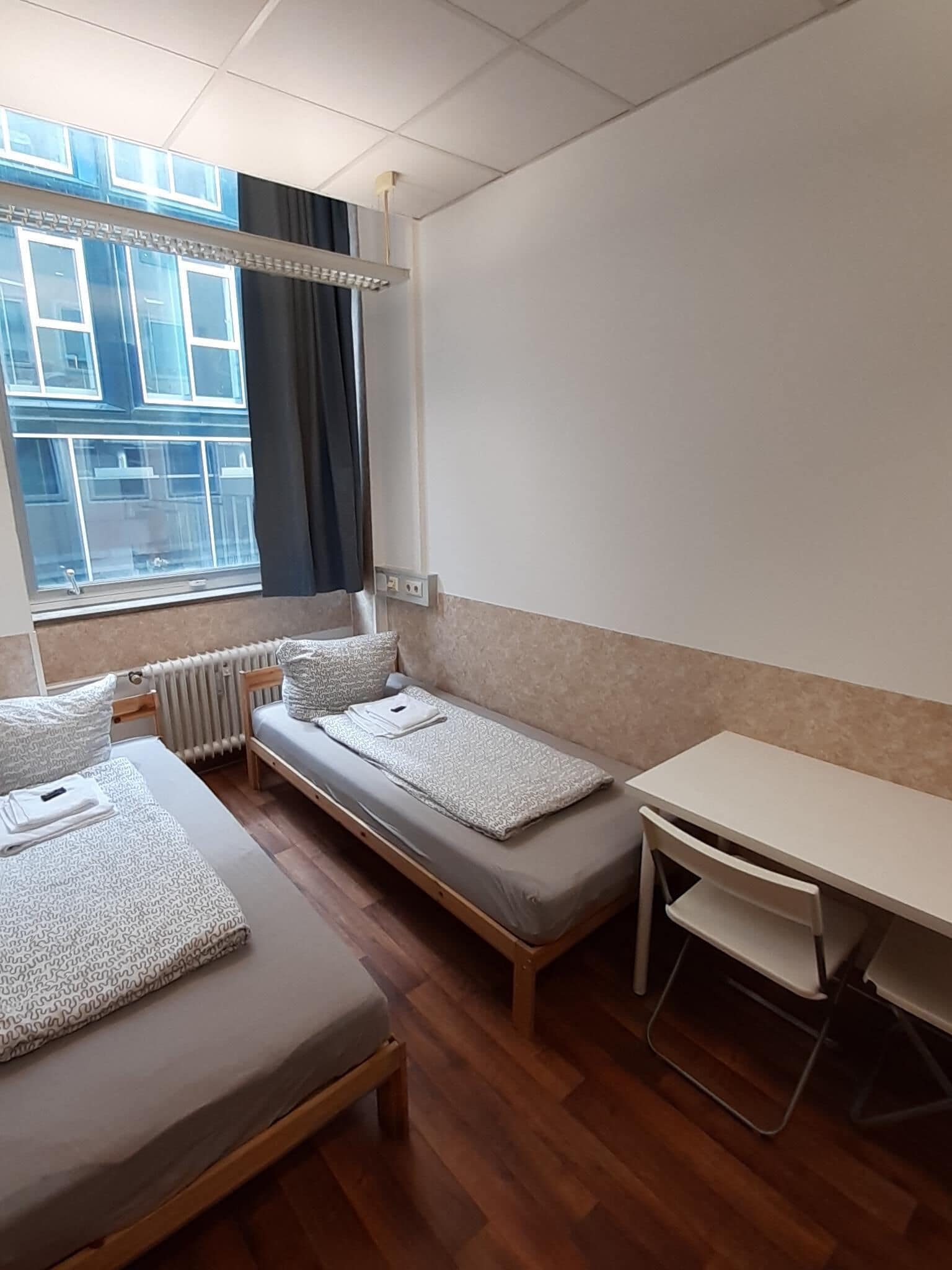 Quarto pequeno e compacto com duas camas de solteiro, uma escrivaninha e uma janela com cortinas.