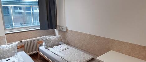 Quarto pequeno e compacto com duas camas de solteiro, uma escrivaninha e uma janela com cortinas.