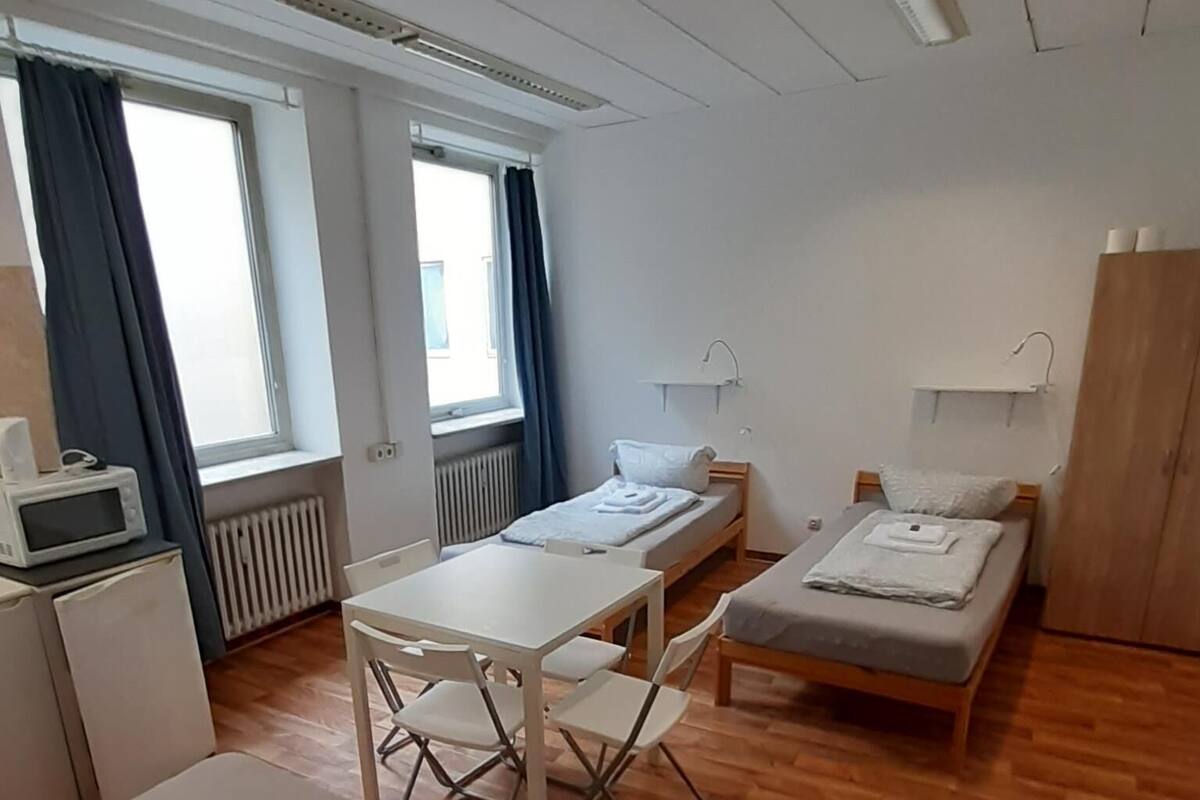 A small, clean room with a wooden floor, a bed, a desk, and a chair. There is a window with curtains, a radiator, and a small table with a microwave.