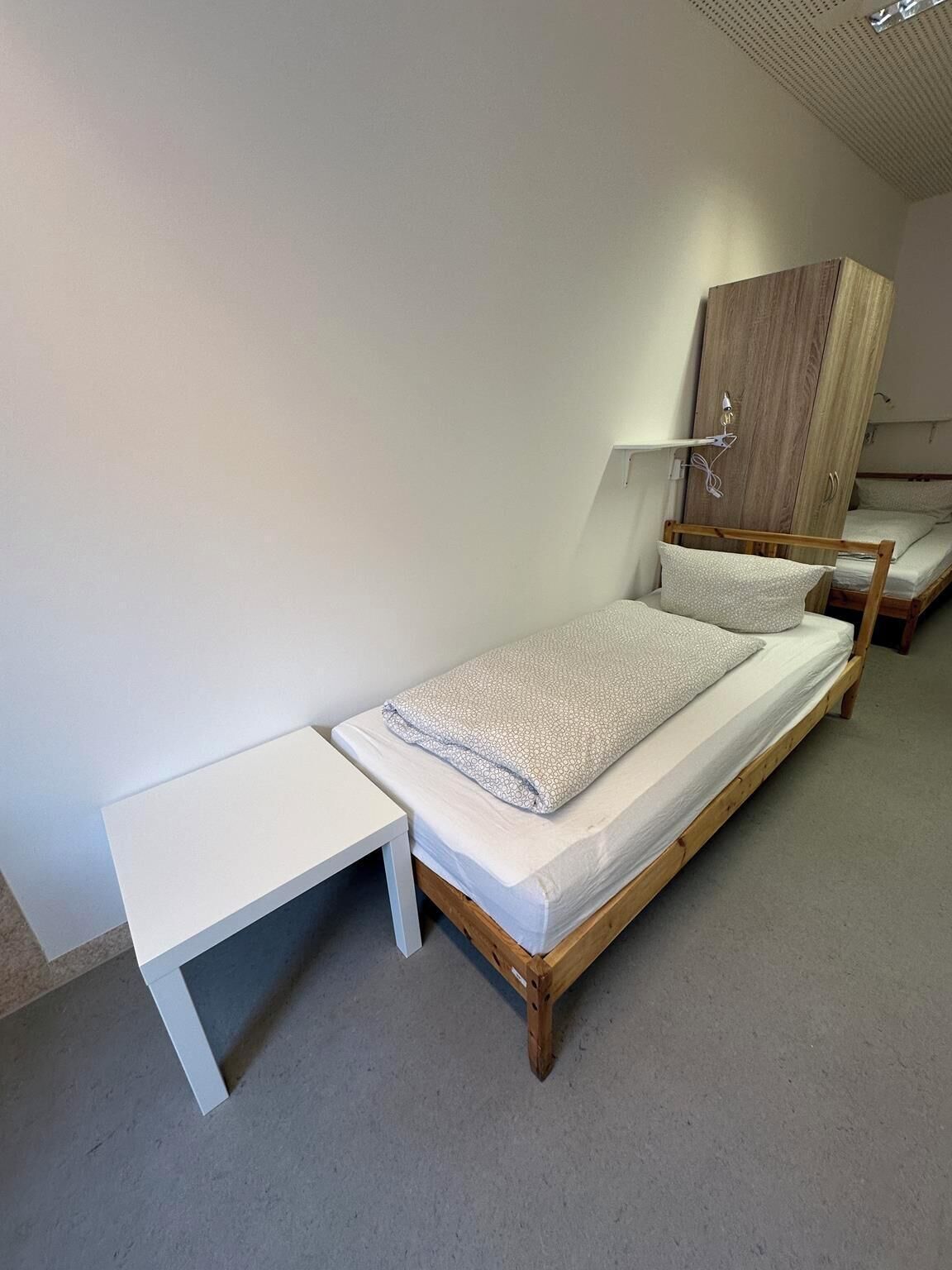 Ein Einzelbett mit Holzrahmen und weißer Matratze, ein kleiner Nachttisch in Weiß sowie ein Holzschrank mit Spiegel.