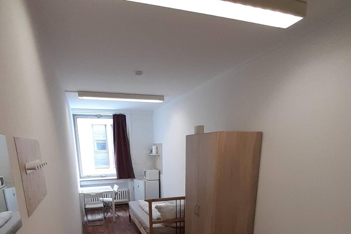 A narrow room with a wooden floor, a bunk bed, a small table with chairs, a radiator, and a window with curtains.