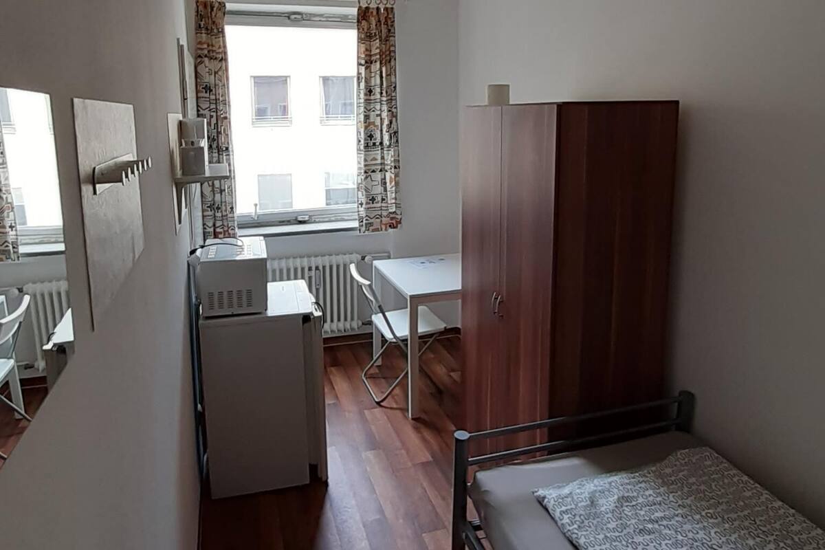 A small, well-lit room with a bed, a desk, and a wardrobe. There is a radiator under the window and a ceiling light.