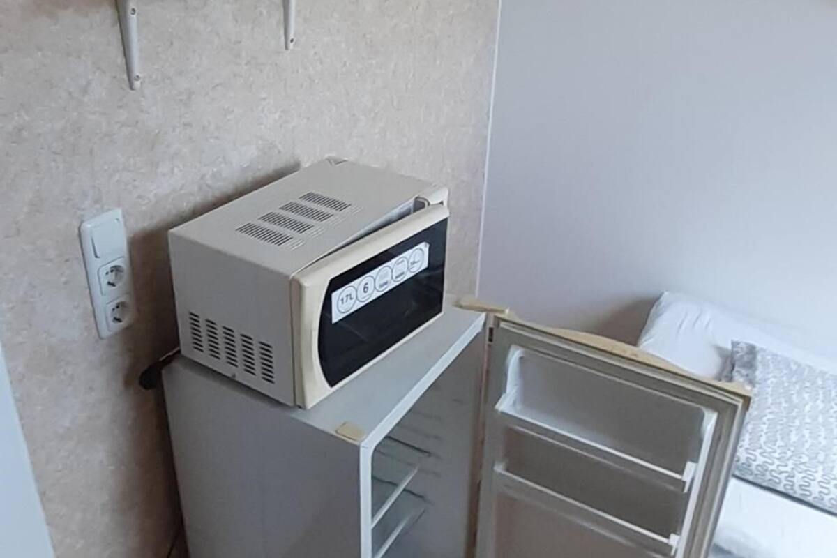 A compact kitchenette with a microwave, a small refrigerator, and a shelf above holding a kettle and glasses.