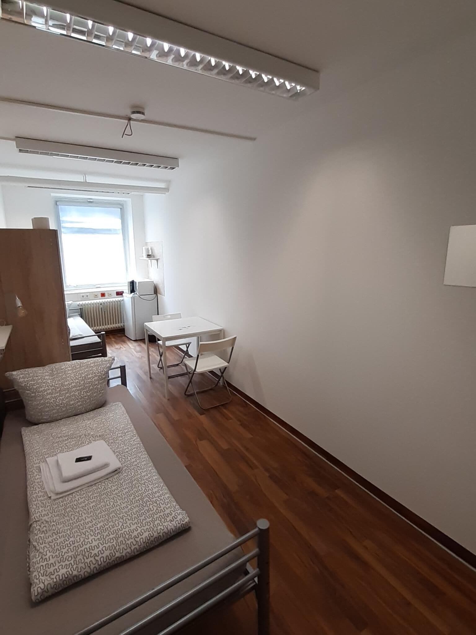 Quarto moderno e bem iluminado, com piso de madeira, um sofá branco com almofadas estampadas, uma pequena mesa e uma cadeira.