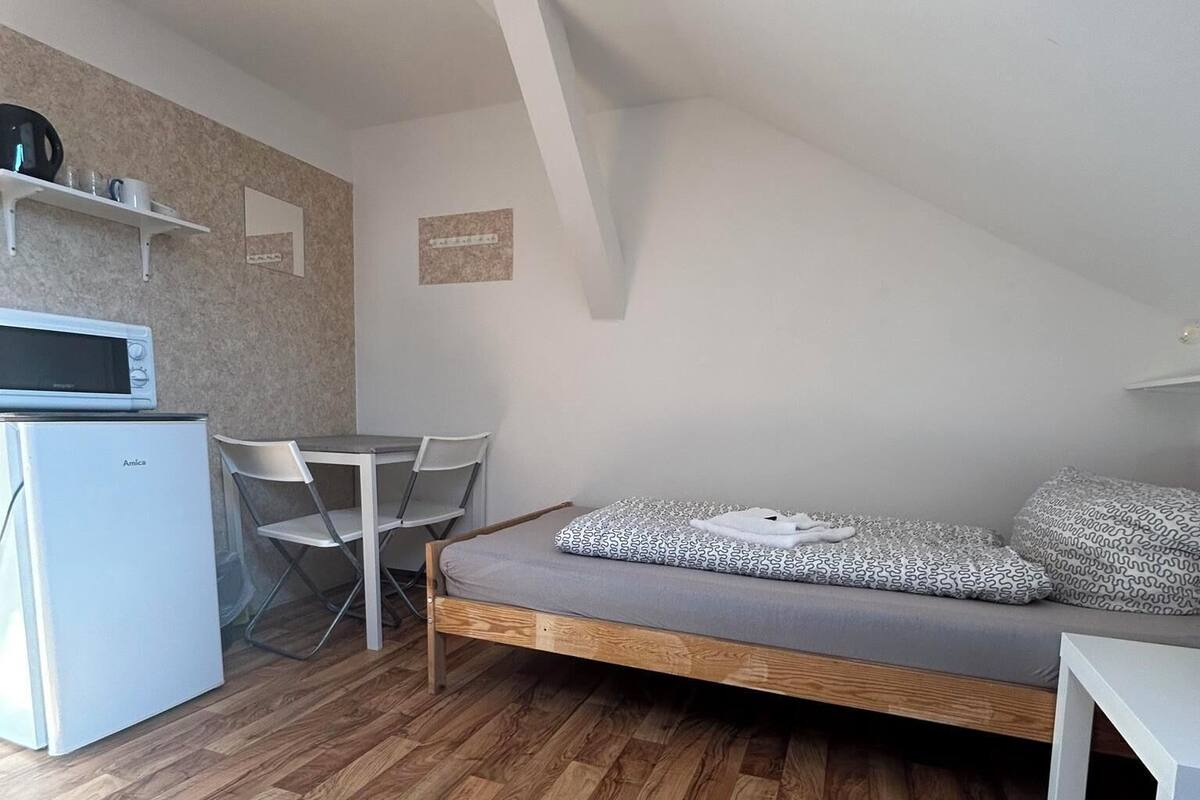 A compact room with a bed, a small table, a chair, a microwave, and a shelf with cups and a kettle.