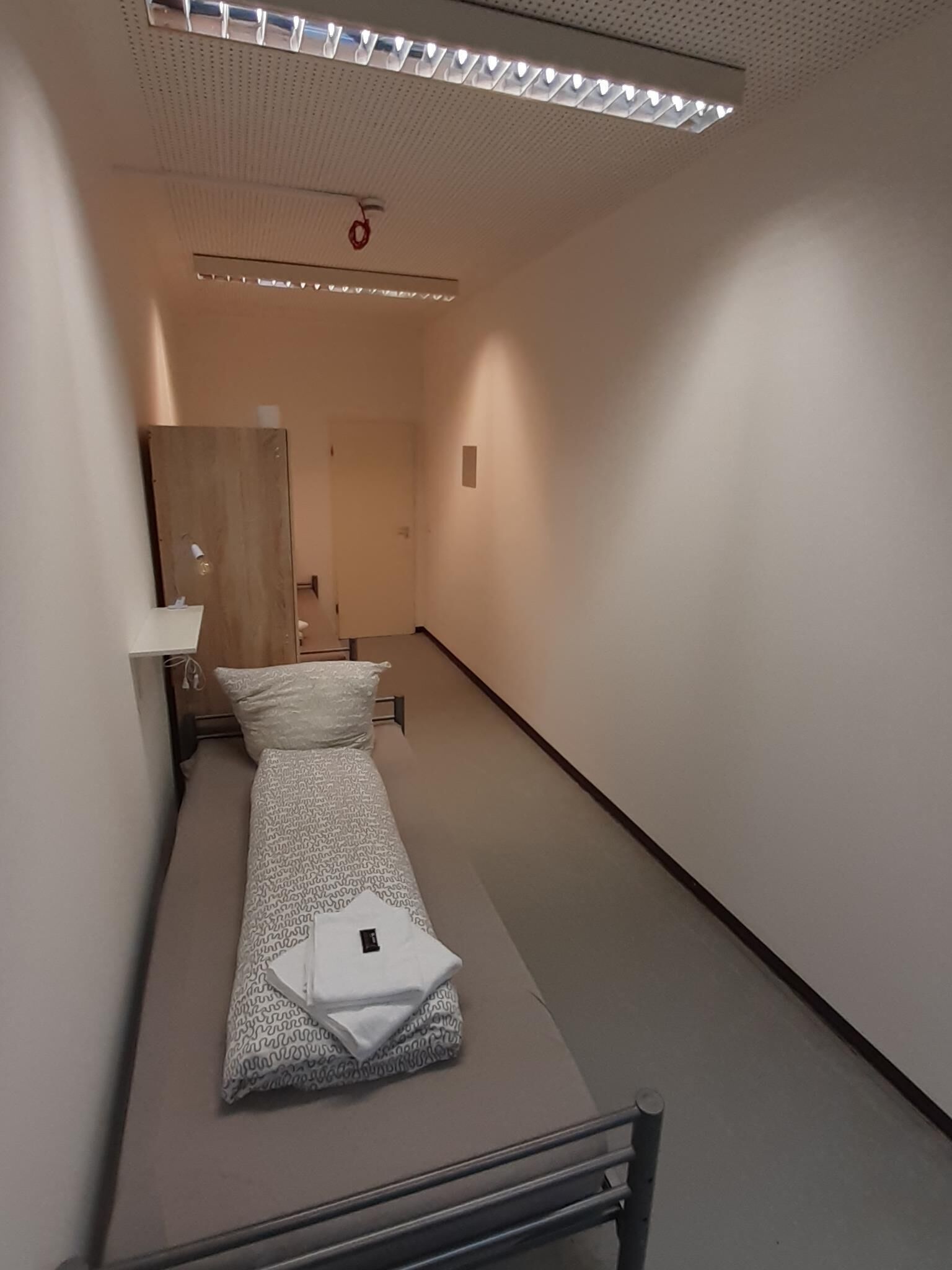 A narrow, dimly lit hallway with a single bed on the left, a wooden door at the end, and a ceiling light fixture.