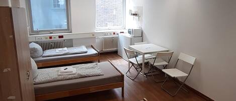 A compact, well-lit room with two bunk beds, a small table with chairs, a radiator, and a window providing natural light.