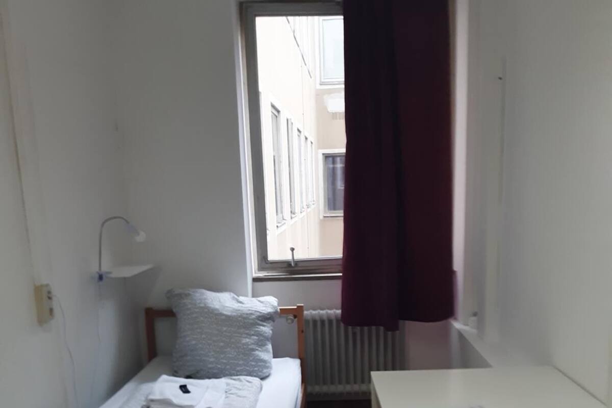 A small, single-bed room with a wooden bed, a white desk, and a chair. There is a window with a curtain, a radiator, and a wall-mounted lamp.