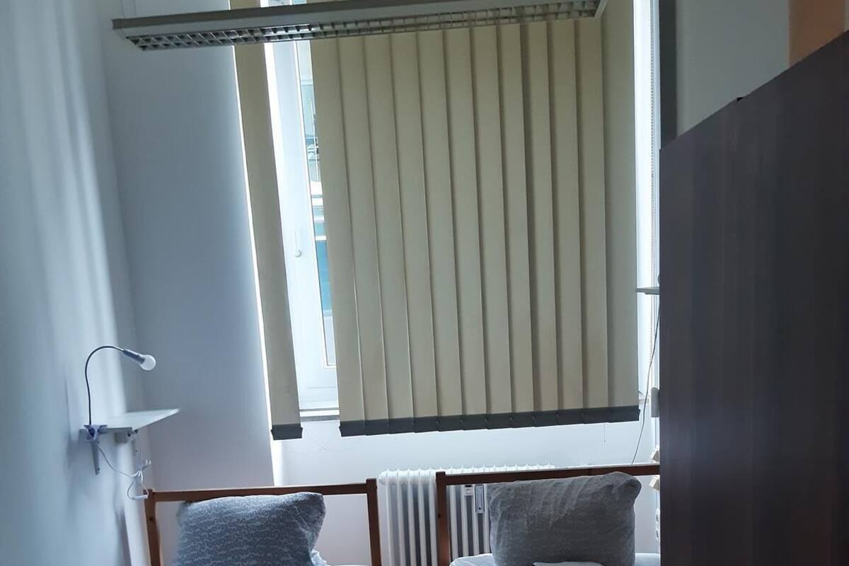 A small, clean room with two single beds, a radiator, a window with blinds, and a wooden wardrobe.