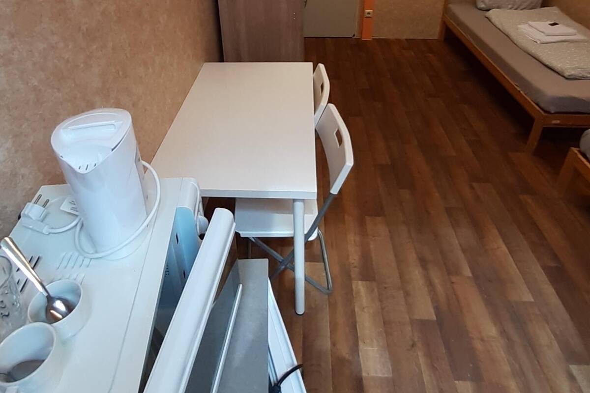 A compact room with a white table and chairs, a bed with a grey mattress, and a wooden wardrobe.
