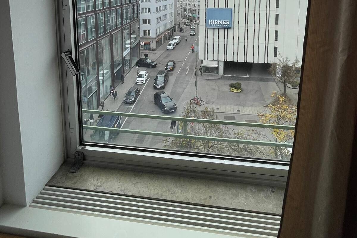 A view from a window overlooking a street with buildings, including a modern structure with a sign that reads "HIRMER REITENMODEL".