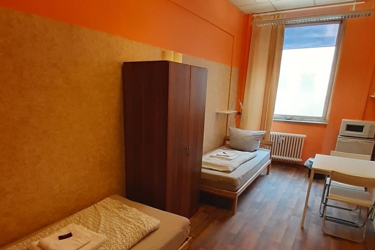 A room with a wooden floor, a bed with a beige mattress and patterned bedding, a wooden wardrobe, a small table with a chair, and a window with curtains.