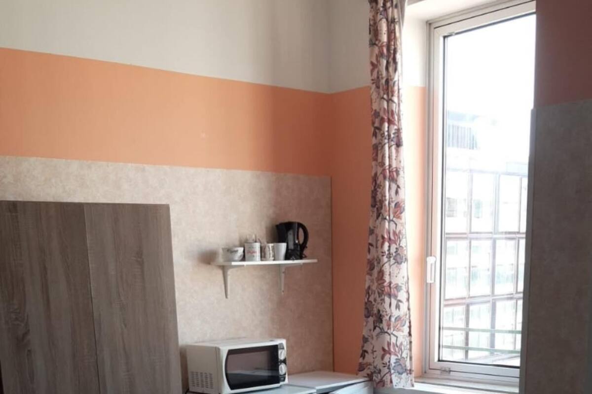 A compact kitchen with a microwave, a small refrigerator, and a window with curtains. There is a table with chairs and a cabinet with doors.