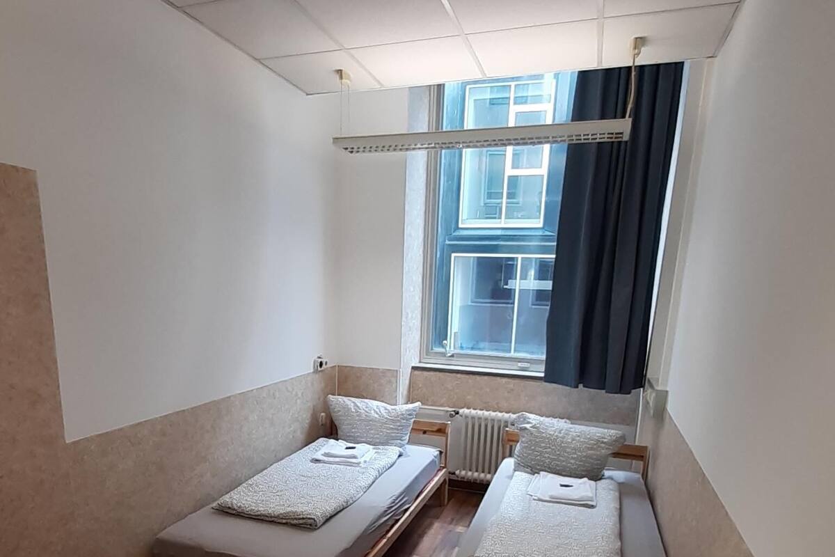 A small, well-lit room with a bed, a table, and a window with curtains.