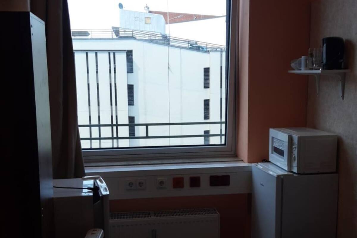 A hotel room with a bed, a desk, a radiator, a window with a view of buildings, and a small shelf with items.