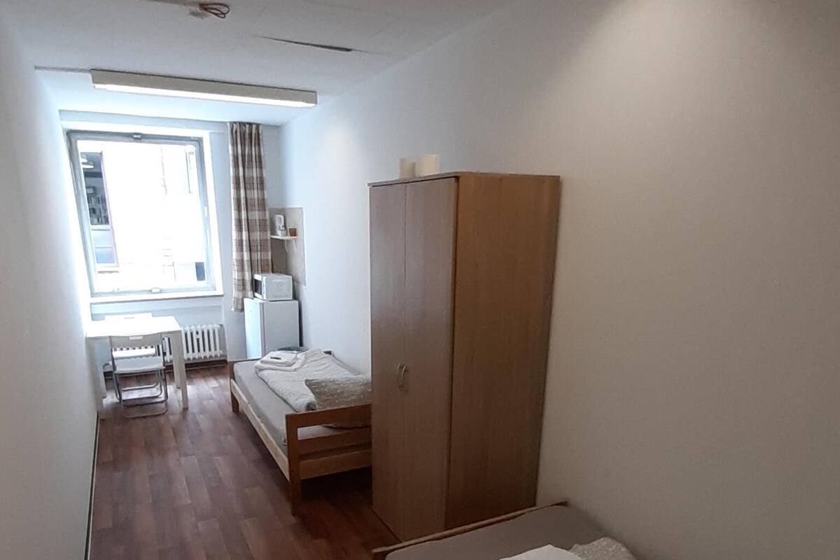 A small, sparsely furnished room with a single bed, a wooden wardrobe, a desk with a chair, and a radiator.