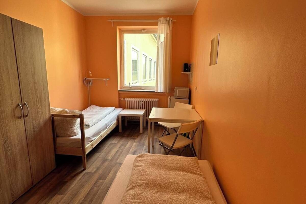 A compact room with two single beds, a small table, a chair, a wardrobe, and a window with curtains.