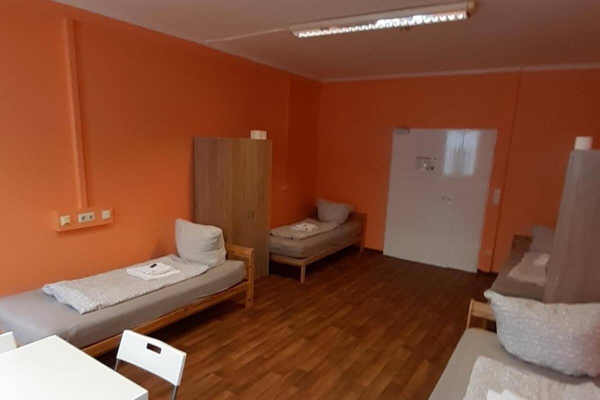 A small, orange-walled room with a wooden floor, a white table with chairs, a bed, a wardrobe, and a door.
