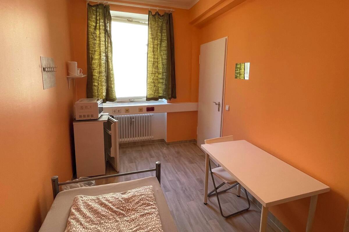 A small, compact room with a window, a radiator, a desk, and a bed.