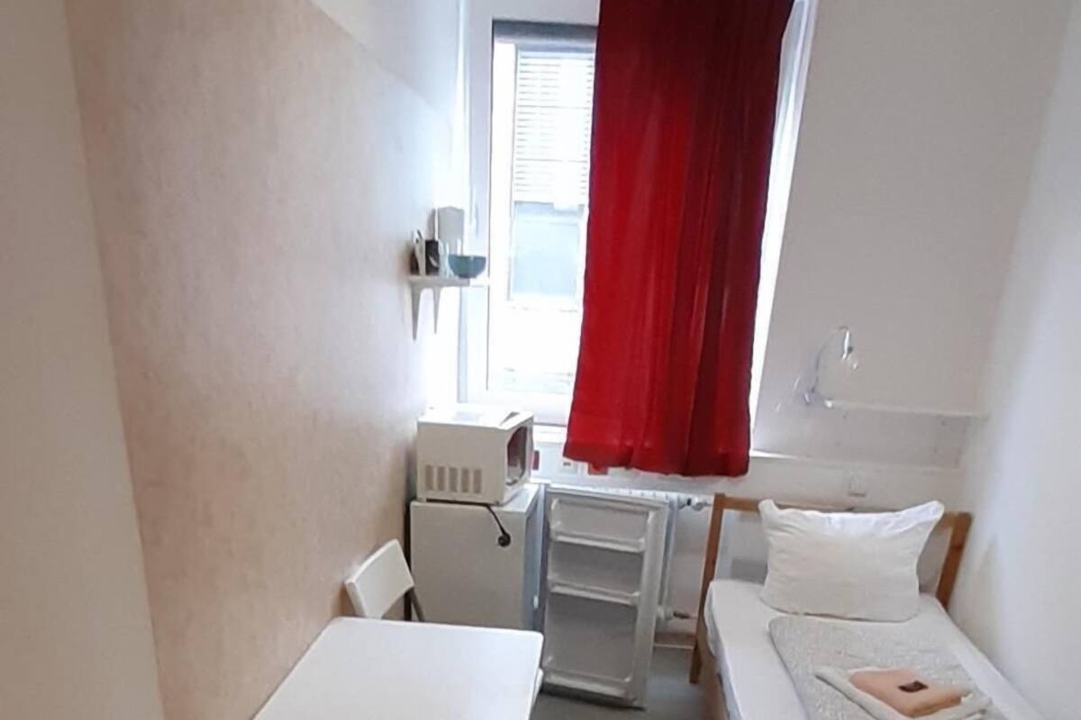 A compact room with a bed, a small table, a chair, a radiator, and a window with red curtains.