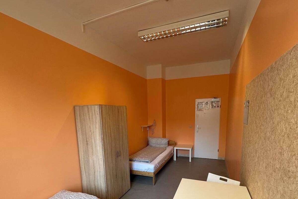 A small, compact room with two single beds, a wooden wardrobe, a small table with a chair, and a whiteboard on the wall.