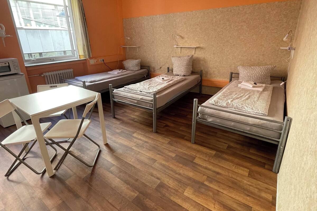A dormitory room with two single beds, a small table, chairs, a radiator, and a window.