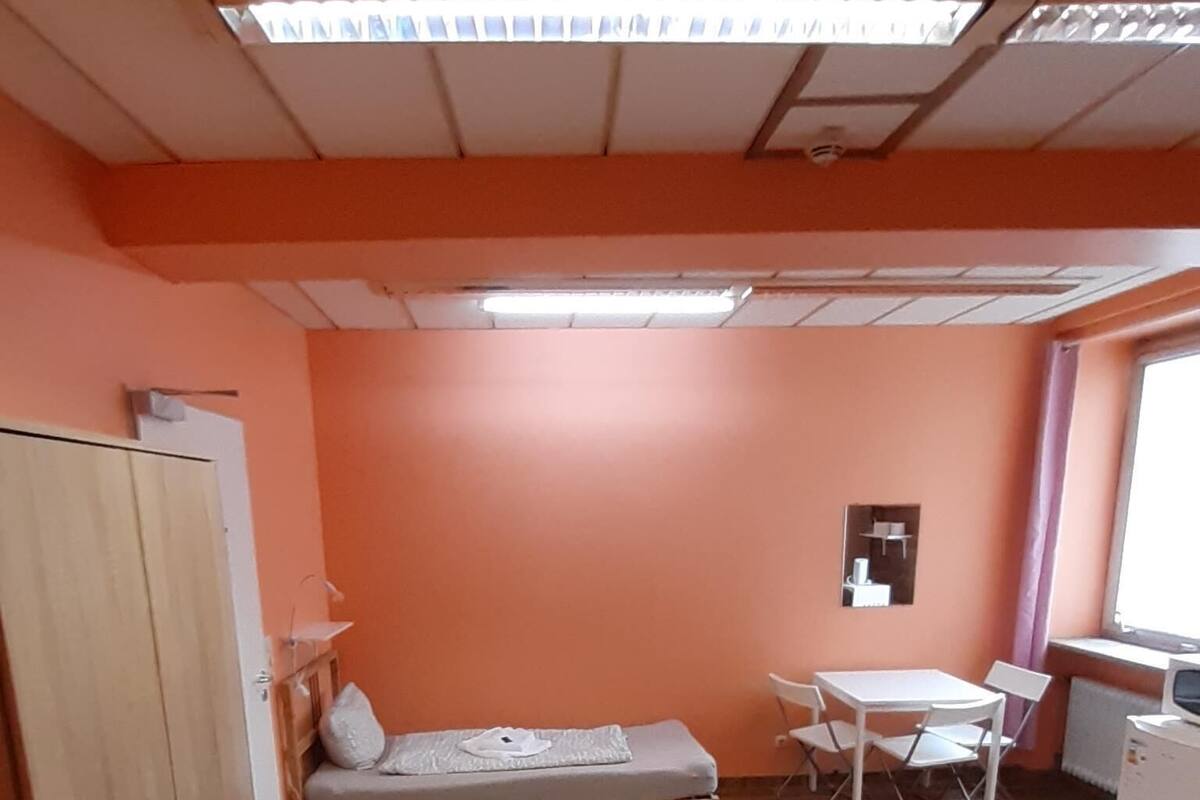 A room with a wooden wardrobe, a single bed, a small table with chairs, a window, and a wall-mounted mirror.