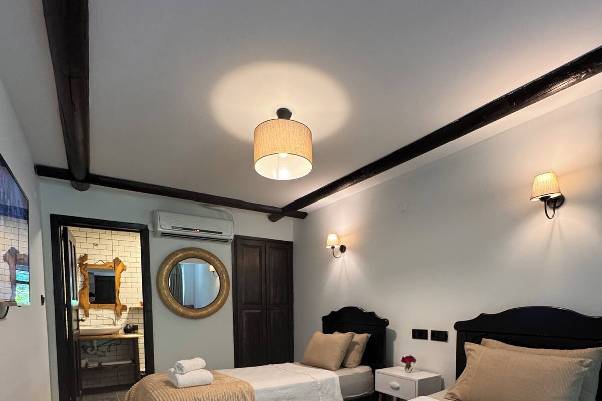 Una habitación de hotel con dos camas, un espejo grande, un espejo redondo, una mesita de noche con una lámpara y una lámpara de pared.
